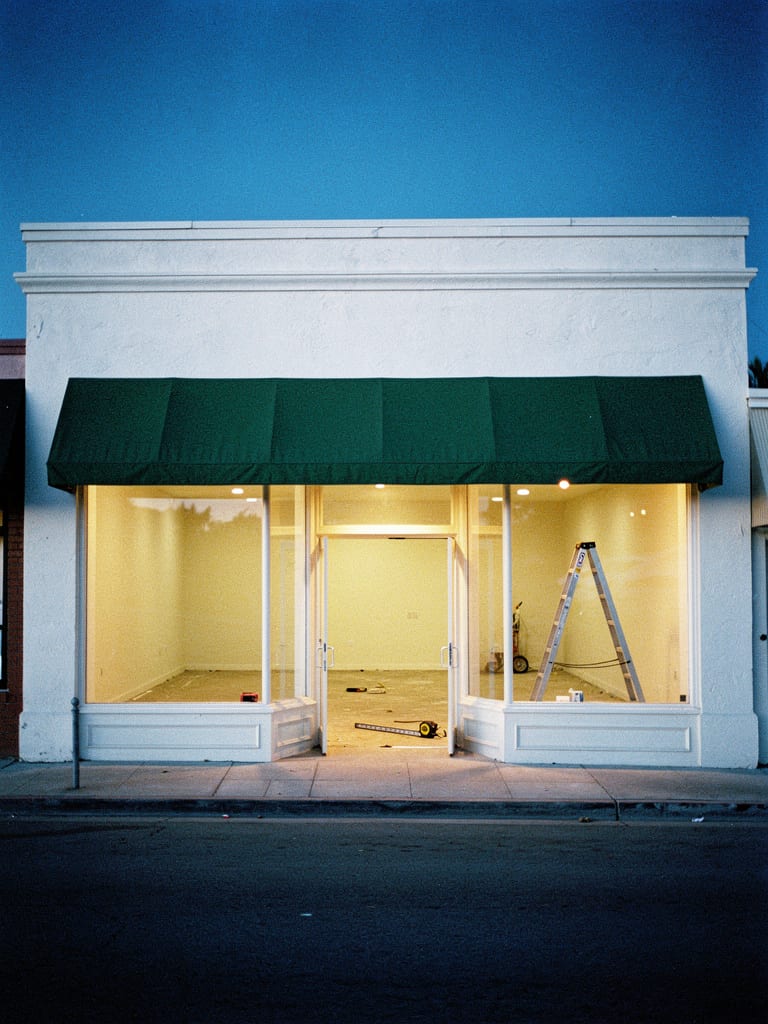A small vacant Lafayette commercial storefront at dusk with warm interior light through its plain glass windows